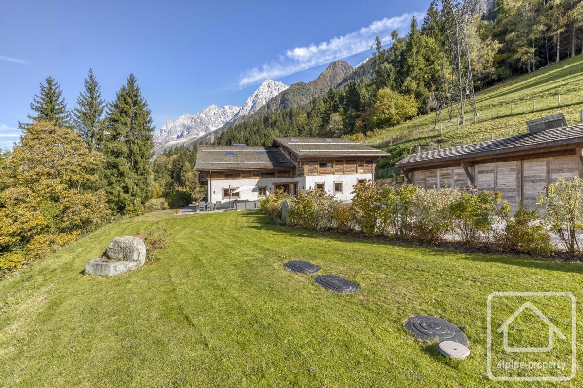 Chalet with a green lawn, surrounded by evergreen trees and majestic snow-capped mountains under a clear blue sky.