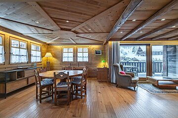 A cozy, rustic wooden chalet interior features a dining table, armchair, and large windows overlooking a snowy landscape.