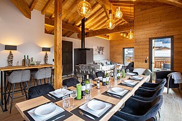 Cozy, modern wooden chalet dining area featuring a long set table under exposed beams and warm hanging pendant lights.
