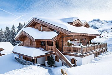 A large, ornate wooden chalet covered in deep snow, nestled in a sunny winter mountain landscape with ski slopes visible in the distance.