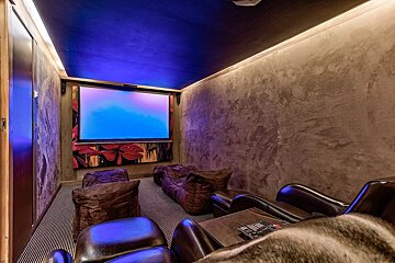 A cozy home theater with a large screen displaying blue hues, a dark blue ceiling, textured walls, and various brown seating, all lit by ambient lights.