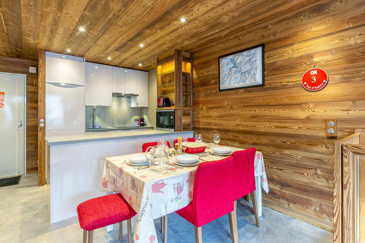 A modern kitchen and dining area with white units, red chairs, and warm wooden walls/ceiling, creating a cozy yet contemporary space.