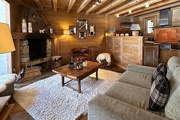 A cozy, rustic chalet living room with wood-paneled walls, a stone fireplace, plush rug, comfortable sofa, and a compact kitchen area.