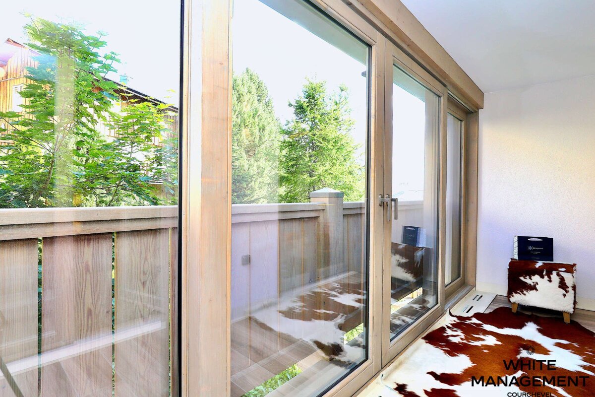 A bright room features large glass doors opening to a wooden balcony with lush green trees. Cowhide rugs and decor add a rustic touch inside.