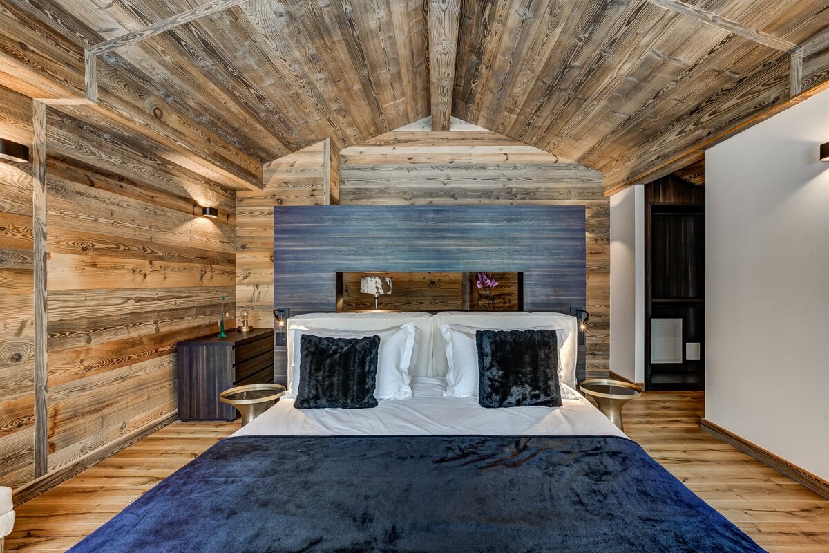 A luxurious chalet-style bedroom featuring warm reclaimed wood walls and a vaulted ceiling. A large bed with a blue headboard and dark bedding completes the cozy space.
