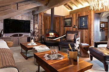 A luxurious, cozy living room with wood paneling, exposed beams, a modern hanging fireplace, large TV, and plush seating, complete with champagne.