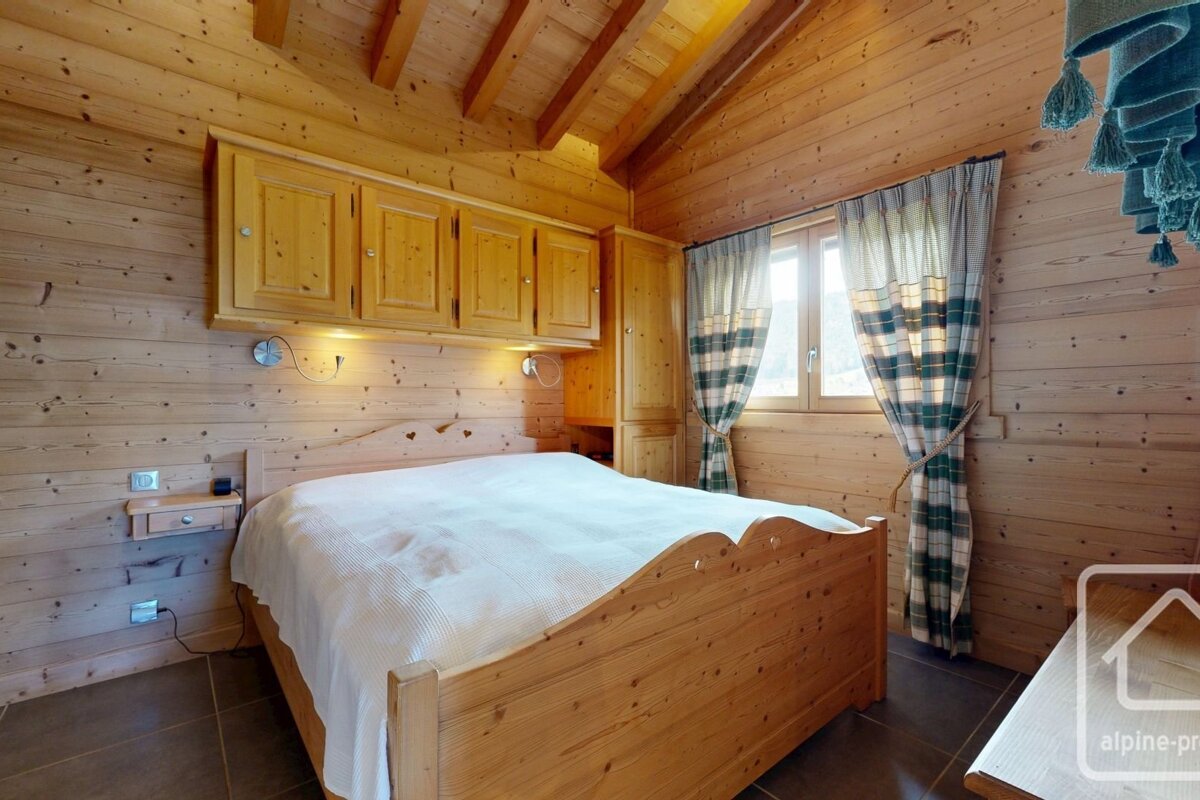 A warm, rustic bedroom with full wood paneling, a large wooden bed, built-in cabinets, and a window with plaid curtains.