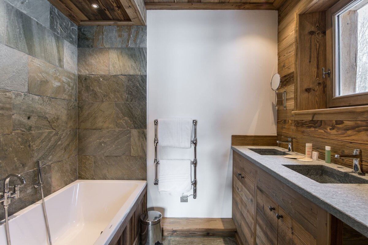 A modern rustic bathroom features a bathtub next to stone-tiled walls, a chrome towel warmer, and a double wooden vanity with stone sinks.