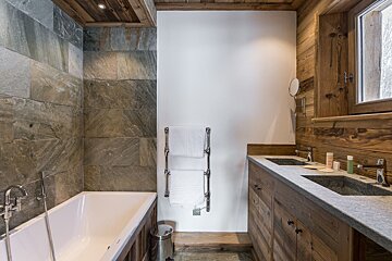 A modern rustic bathroom features a bathtub next to stone-tiled walls, a chrome towel warmer, and a double wooden vanity with stone sinks.