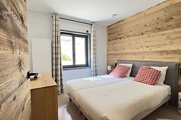 A bright bedroom with two single beds, white duvets, and red plaid pillows. It features a rustic wooden accent wall and a window with plaid curtains.