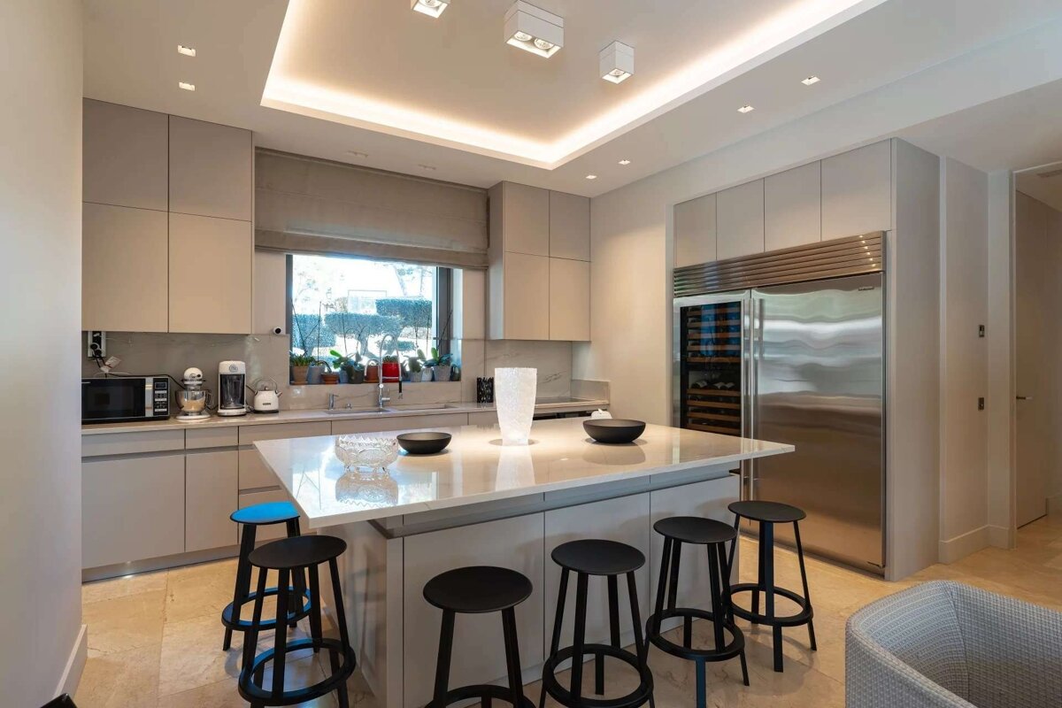 A bright, modern kitchen features light gray cabinets, a large island with black stools, stainless steel fridge/wine cooler, and a window with plants.