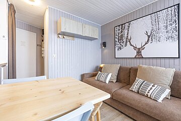 Cozy, modern cabin living room with light plank walls, a brown sofa, and a large winter deer art. A wooden dining table is in the foreground.