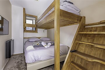 A compact bedroom features wooden bunk beds with a larger bottom mattress, wooden stairs, and a wall-mounted TV.