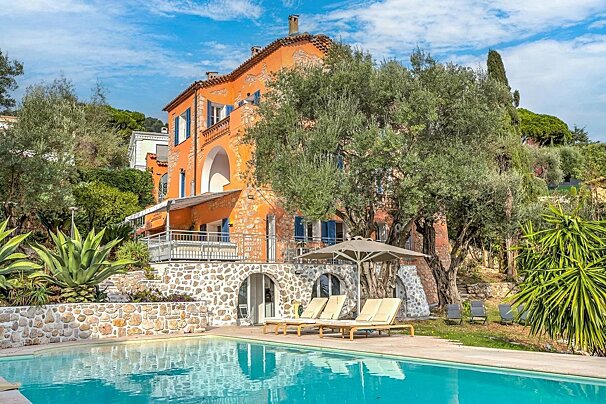 A sunny Mediterranean villa with an orange exterior, blue shutters, a swimming pool, lounge chairs, and lush olive trees.