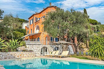 A sunny Mediterranean villa with an orange exterior, blue shutters, a swimming pool, lounge chairs, and lush olive trees.