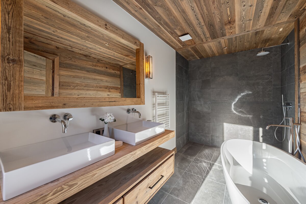 Chic rustic bathroom featuring a wooden ceiling, vanity with double white sinks, grey tiled shower, and a sleek freestanding tub.