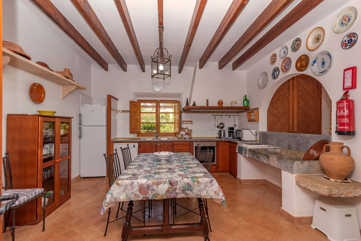 A rustic kitchen with wooden ceiling beams, a dining table, and traditional decor. It features a stone sink, white fridge, and window.