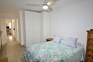 A modern bedroom with white walls, a wood ceiling fan, large white wardrobes, and a bed with a green patterned cover.