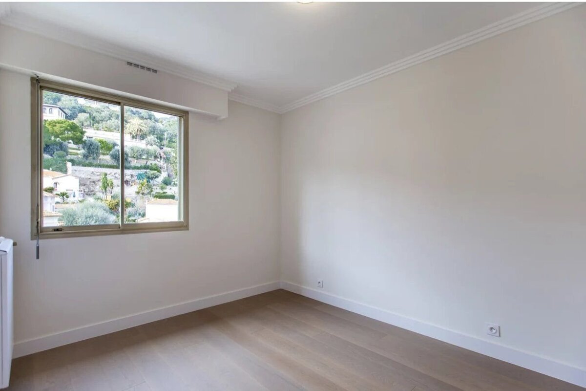 An empty room featuring light wood floors, white walls, and a window with a scenic view of a green hillside.