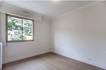 An empty room featuring light wood floors, white walls, and a window with a scenic view of a green hillside.