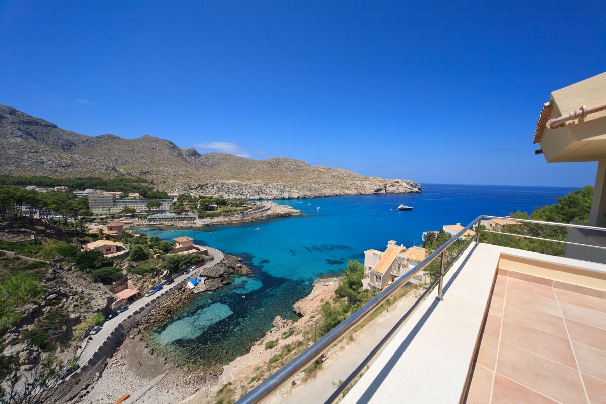 Panoramic view of a vibrant turquoise bay with clear water, surrounded by coastal buildings and rocky mountains, all under a brilliant blue sky, from a balcony.
