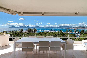Balcony with a white dining set overlooking a marina full of yachts, the blue sea, distant mountains, and palm trees under a bright sky.