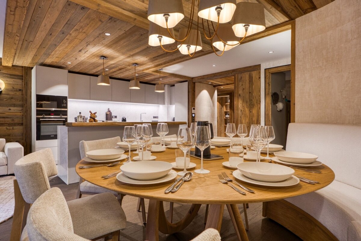 Modern dining area with a rustic wooden ceiling and a round table set for guests, blending into a contemporary kitchen with light cabinets.