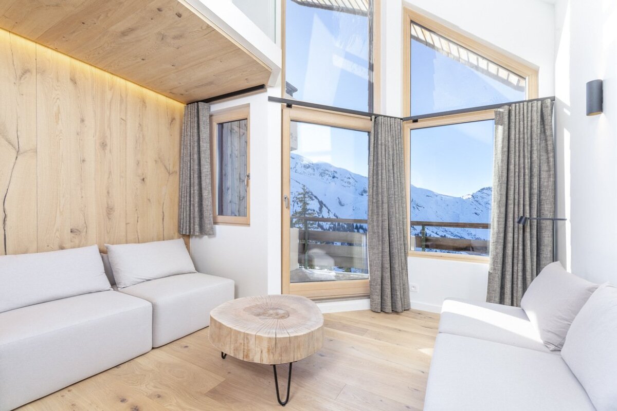 Modern chalet living room with light-colored sofas, a log coffee table, and large windows showcasing snowy mountain peaks.