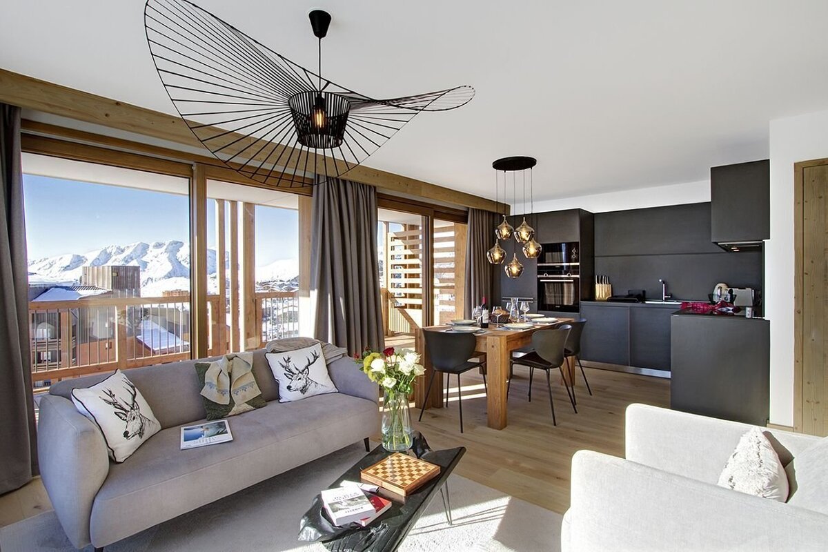 Modern ski chalet living area with a dark kitchen, dining space, and gray sofa. Floor-to-ceiling windows offer a spectacular view of snow-capped mountains.