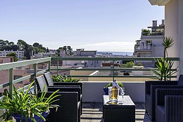 Sunny balcony terrace with wicker furniture, potted plants, and a champagne bucket, overlooking a cityscape and the sparkling sea.