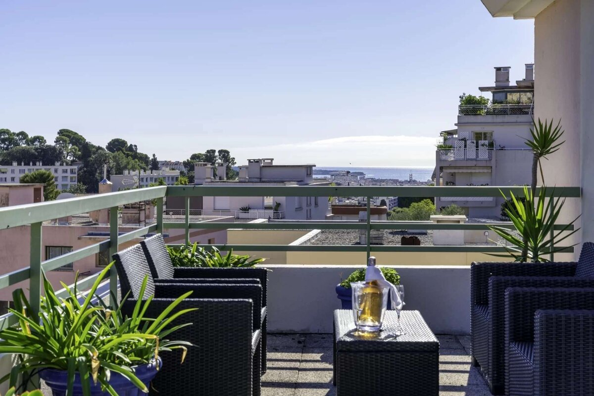 A bright balcony with outdoor seating and drinks, overlooking a city and the sea under a clear blue sky.