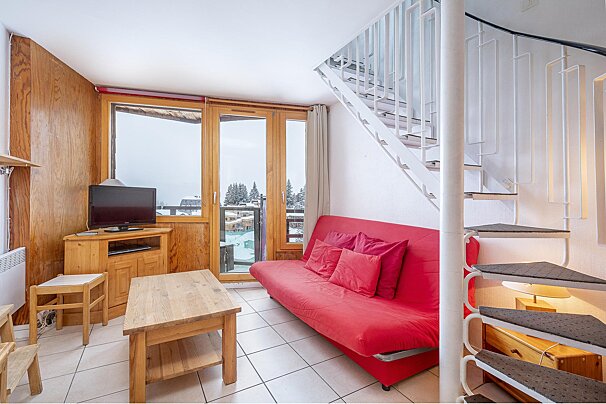 A cozy living room with a red sofa, wooden furniture, a TV, and a spiral staircase. A large window offers a snowy mountain view.