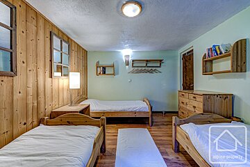 Rustic bedroom with three single wooden beds, wood-paneled wall, light green walls, wooden floor, dresser, and shelves.