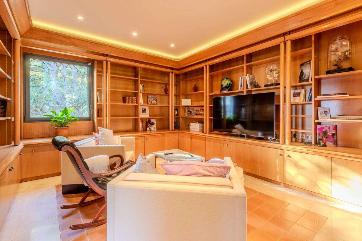 A cozy wood-paneled room with built-in bookshelves, a TV, comfy sofas, a rocking chair, and a window with natural light.