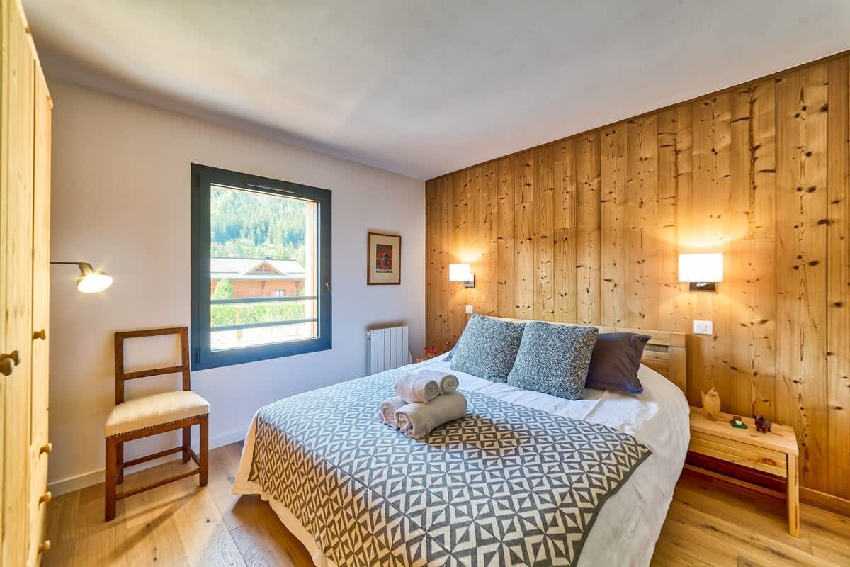 Cozy rustic bedroom with a wood-paneled wall, a double bed with patterned linens, a wooden chair, and a window showing scenic views.