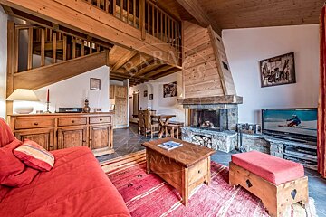 Rustic chalet living room with a stone fireplace, wooden beams, red sofa, and a loft. A dining area and TV with a skier are also visible.