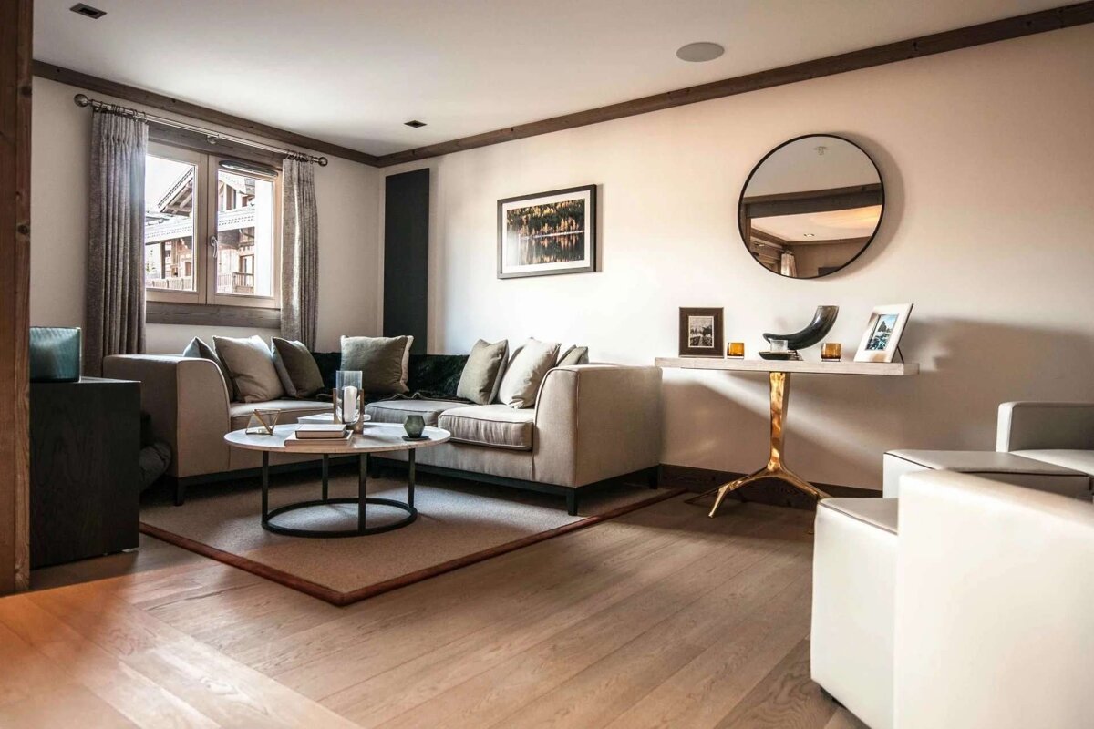 A modern, upscale living room with a large L-shaped couch, coffee table on a rug, wood floors, and a window with curtains.
