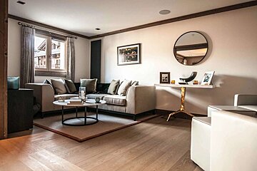 A modern, upscale living room with a large L-shaped couch, coffee table on a rug, wood floors, and a window with curtains.