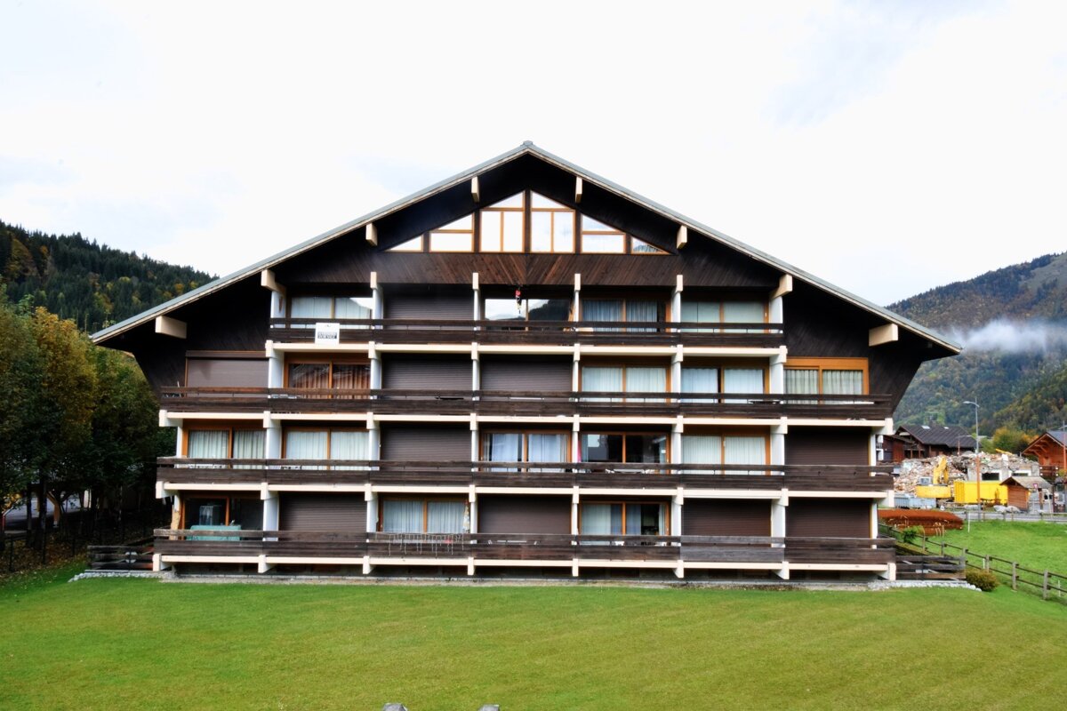 A multi-story wooden alpine building with balconies, situated on a green lawn against a mountain backdrop.