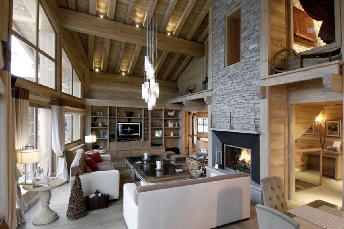 A luxurious, cozy chalet living room with high beamed ceilings, large windows, a stone fireplace, modern furniture, and warm lighting.