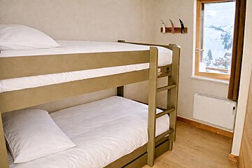 A cozy room features olive green bunk beds with white bedding, a window overlooking a snowy mountain landscape, and ski-themed decor on the wall.