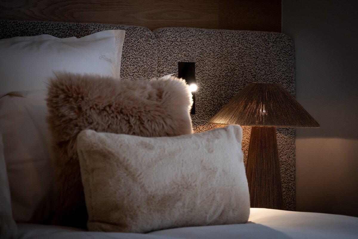 Cozy bedroom scene with plush furry pillows on a bed, illuminated by a warm table lamp and a small light on a textured headboard.