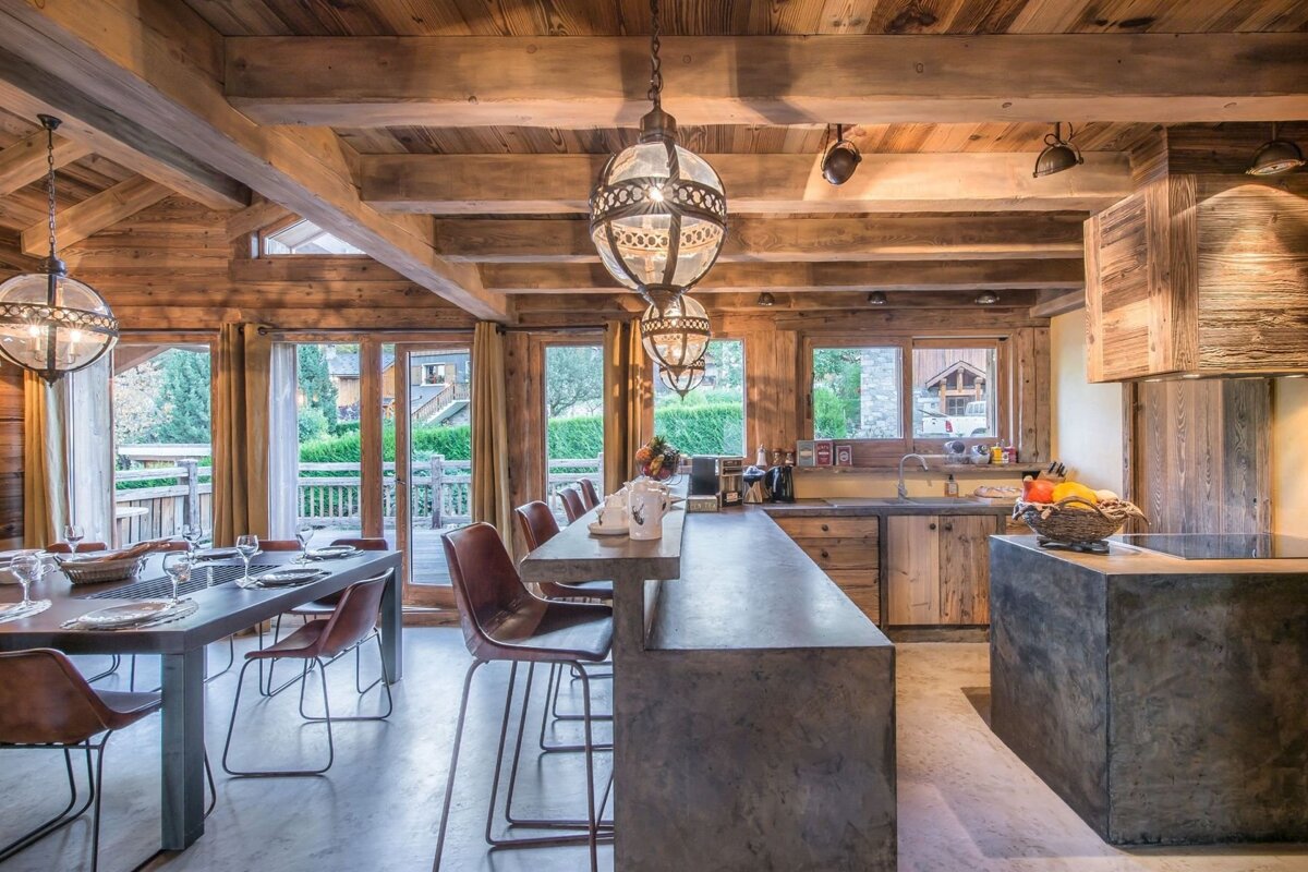A luxurious, open-plan rustic lodge interior with a wooden beam ceiling, modern kitchen with concrete counters, dining area, and large windows looking outdoors.