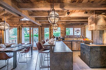 A luxurious, open-plan rustic lodge interior with a wooden beam ceiling, modern kitchen with concrete counters, dining area, and large windows looking outdoors.