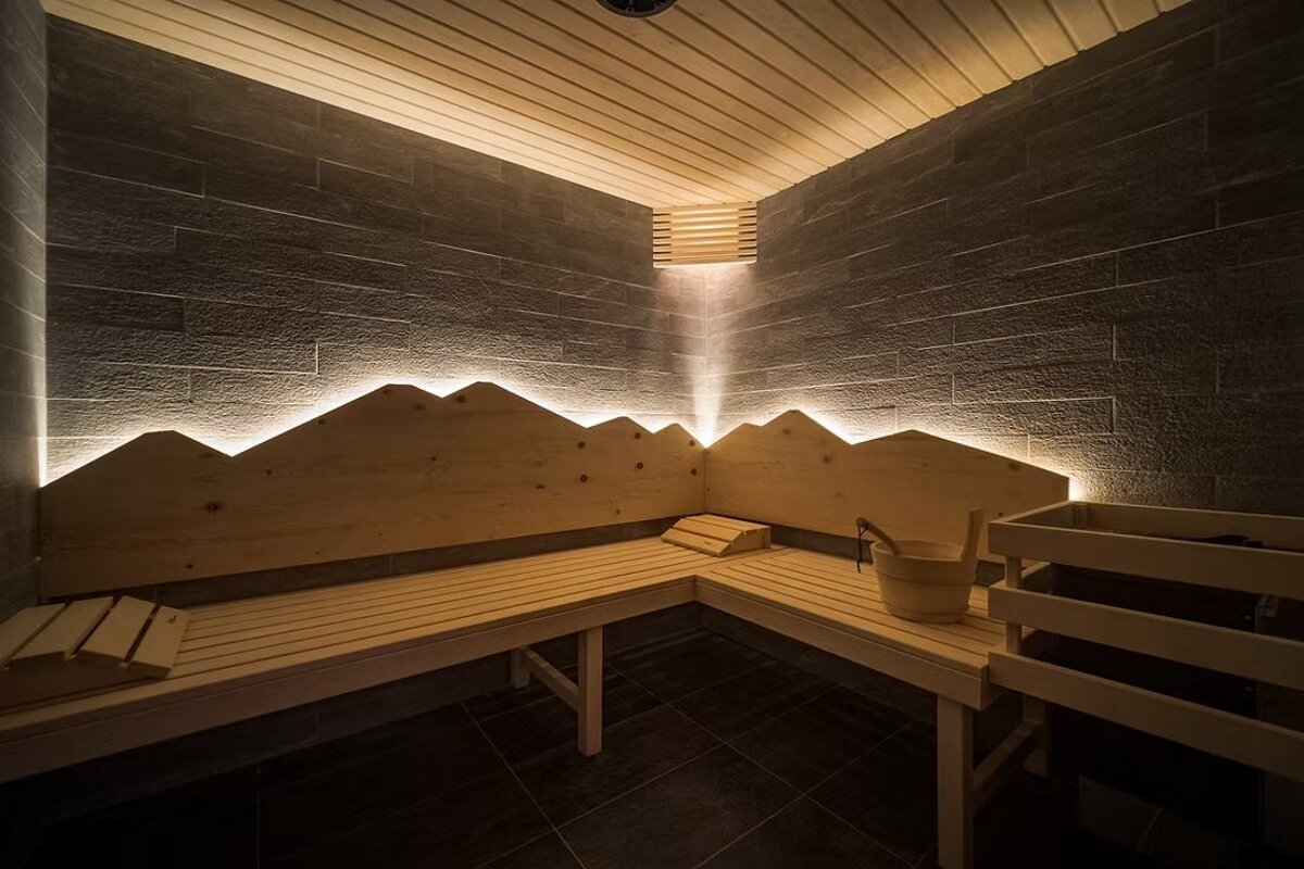 Modern sauna interior featuring L-shaped wooden benches, dark stone walls, and a unique backlit backrest shaped like mountain peaks.