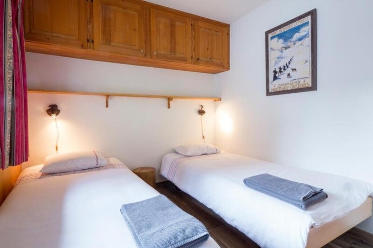 A cozy bedroom featuring two twin beds with white linens, overhead wooden cabinets, and a framed vintage poster on the wall.