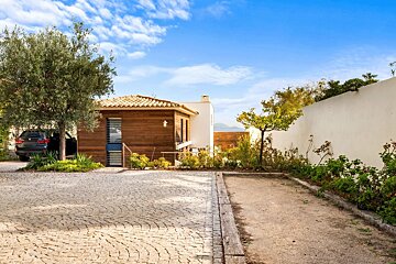 A sunny courtyard with a cobblestone driveway, a wooden cottage, and a pétanque court, under a bright blue sky.
