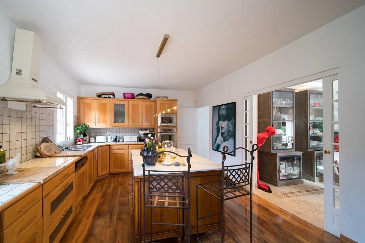 A bright kitchen featuring light wood cabinets, a central island with bar stools, hardwood floors, and a doorway opening to another room.