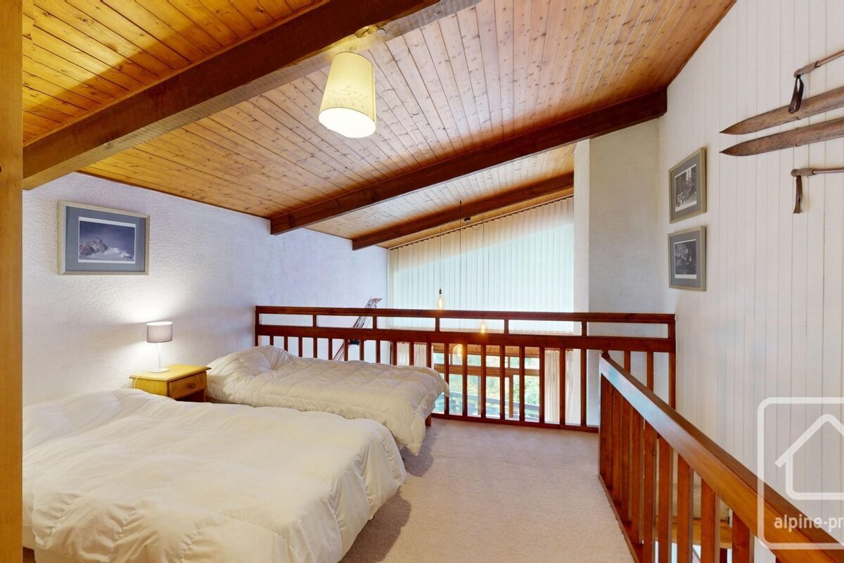 Cozy loft bedroom with two beds, a rustic wooden ceiling, and a wooden railing overlooking a bright living area. White walls with ski decor.