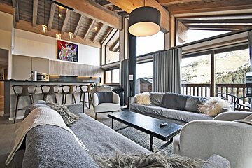 A modern, rustic-chic living room with an open kitchen bar, wooden beams, cozy sofas, and large windows revealing scenic mountain views.